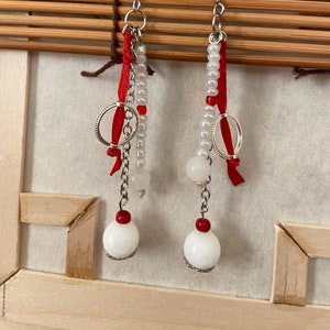 Op de afbeelding: Een paar oorhangers met witte kralen, rood lint en zilveren bedels. De oorhangers hebben een grote witte kraal aan de onderkant.