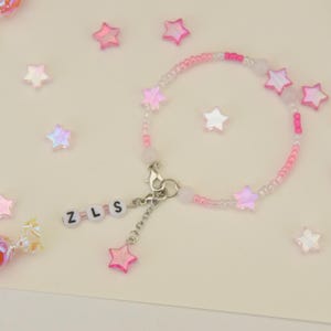 Puede incluir: Un brazalete de cuentas rosa y blanco con un dije de estrella y un dije con las letras "Z-L-S". El brazalete está rodeado de estrellas iridiscentes.