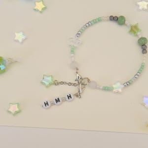 May include: A green and white beaded bracelet with silver accents and star charms. The bracelet has a silver clasp and a charm with the letters "HMMH" on it.