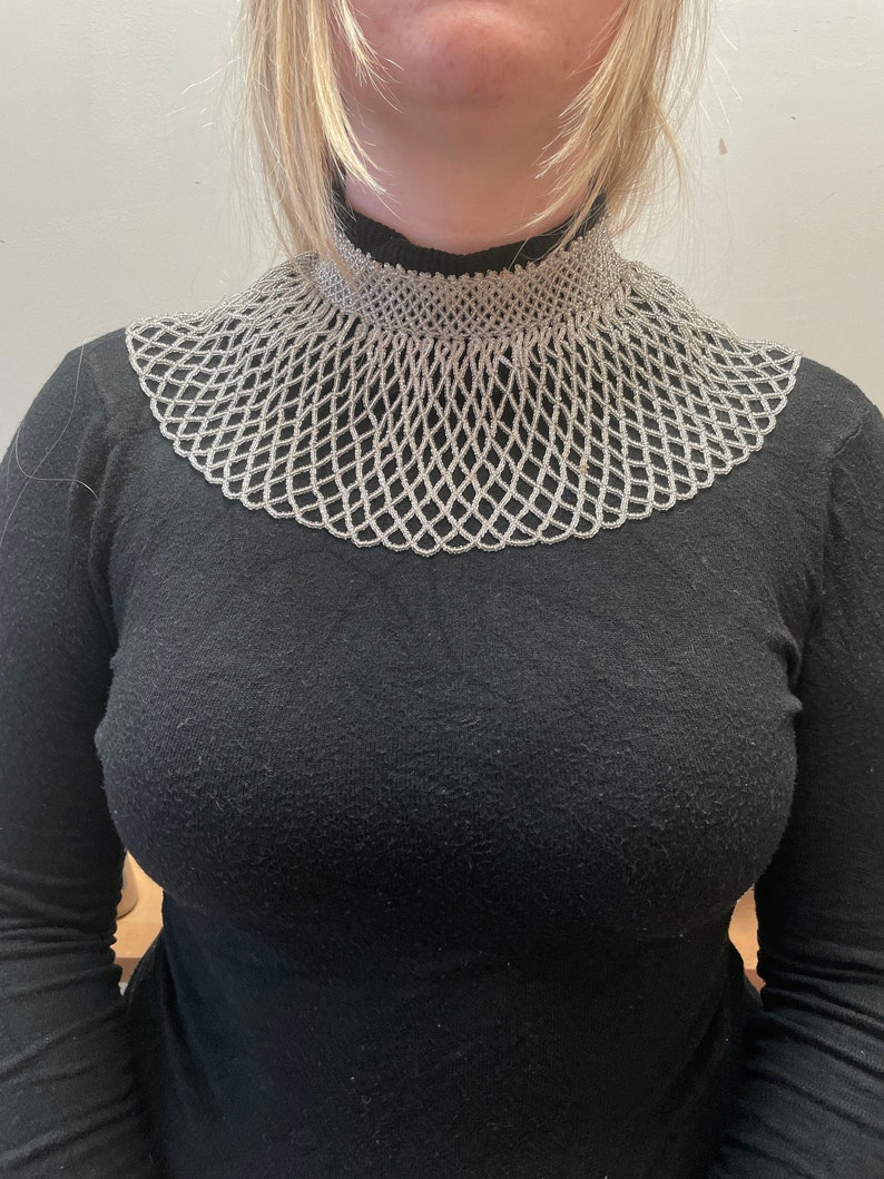 May include: A silver beaded collar necklace with a wide, open design. The necklace is made of small, round beads that are strung together in a delicate pattern.