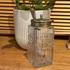 May include: A clear glass sugar dispenser with a silver lid and spoon. The dispenser has a square shape and a decorative pattern.