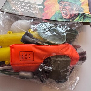 May include: A sealed package of vintage "Combat Man" action figure accessories. The packaging features a screaming soldier illustration and text that reads "30 PC. ACCESSORIES FOR ALL 11 1/2 ACTION FIGURES." The accessories include a yellow tube, an orange vest, and other small items.