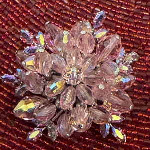 May include: A pink crystal flower brooch with iridescent accents. The brooch features a central cluster of crystals surrounded by petals. The background is a textured, deep red surface.
