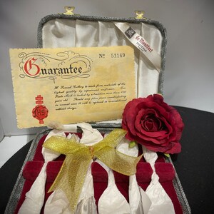 May include: A set of six silver teaspoons in a red velvet lined box with a gold ribbon. The box is closed and has a guarantee card inside. The card has the text "Guarantee" and "No. 51149". A red rose is on top of the box.