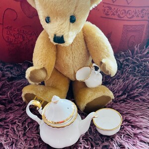 May include: A beige teddy bear holding a white teacup sits on a purple fluffy surface with a white teapot and a white teacup beside it.