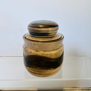 May include: A ceramic jar with a lid. The jar is cylindrical with a cream-coloured base and a dark brown wave pattern. The lid is a rounded shape with a similar colour scheme. The jar is sitting on a clear surface.