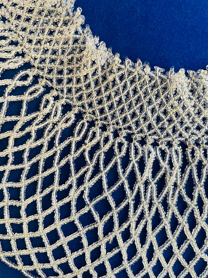 May include: A white beaded collar necklace with a delicate, intricate design. The necklace is made of small, round beads that are woven together to create a lacy pattern.