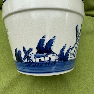 May include: A white ceramic planter with a blue and white design of a windmill, house, and trees. The planter has a crackled glaze.