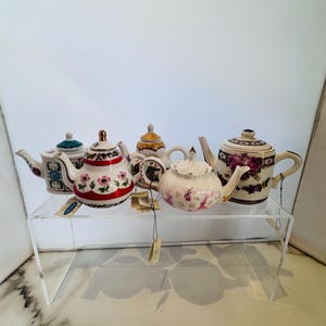 May include: A collection of six decorative teapots with floral designs. The teapots are displayed on a clear acrylic shelf. The teapots feature various colors, including white, red, pink, and gold. Some teapots have intricate patterns and gold accents.