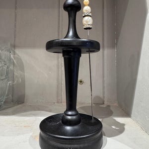 May include: A black wooden pin holder with a decorative pin. The pin has a gold bead, two white beads, and a rhinestone accent. The pin holder has a round base, a central column, and a flat top.