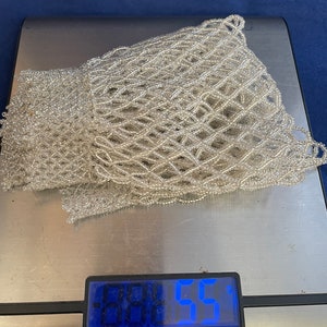 May include: A white beaded net bag on a digital scale that reads 0.0455. The scale is silver and has a black display.