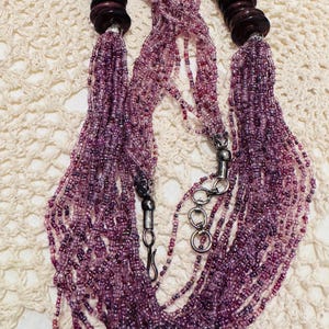 May include: A multi-strand beaded necklace in shades of purple and pink. The necklace features dark brown wooden accents at each end and a silver-toned clasp. The beads are small and create a cascading effect, perfect for adding a touch of elegance.
