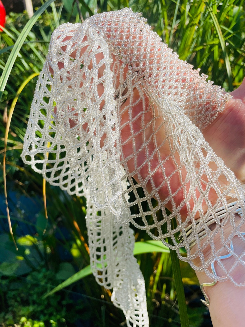 May include: A white beaded scarf with a delicate, intricate design. The scarf is made of small, white beads that are woven together to create a lacy pattern.