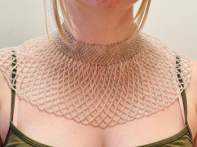 May include: A silver beaded choker necklace with a wide, fan-shaped design. The necklace is made of small, round beads that are woven together in a delicate pattern.