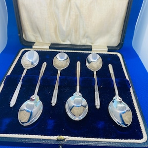 May include: A set of six silver spoons presented in a vintage blue velvet-lined case. The spoons have a polished, reflective surface and decorative handles. The case is rectangular with a black exterior and a cream satin lining.