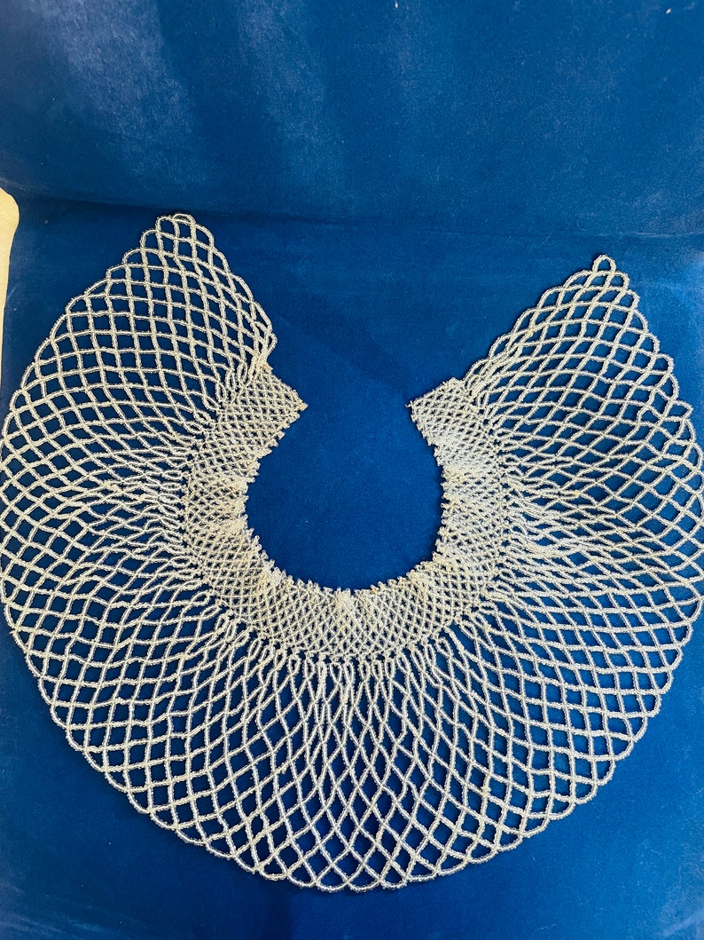 May include: A white beaded collar necklace with a delicate, intricate design. The collar is made of small, white beads woven together in a pattern that resembles a net. The collar is a traditional African accessory.