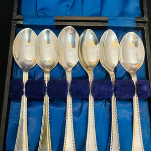 May include: Six silver teaspoons with decorative edges are displayed in a blue velvet lined box. The spoons are arranged in a row, with the handles facing the viewer.