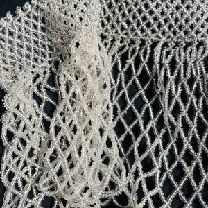 May include: A close-up of a white beaded fringe trim with a diamond pattern. The trim is made of small, clear beads that are strung together to create a delicate and intricate design.