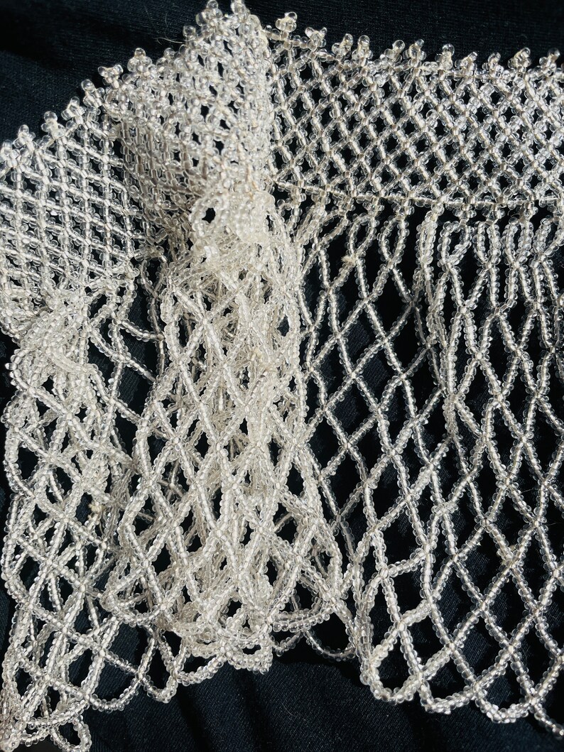 May include: A close-up of a white beaded fringe trim with a diamond pattern. The trim is made of small, clear beads that are strung together to create a delicate and intricate design.