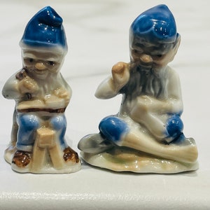 May include: Two small ceramic figurines of gnomes. The gnome on the left is sitting on a stool and holding a hammer. The gnome on the right is sitting on a rock and holding a pickaxe. Both gnomes are wearing blue hats and have white beards.
