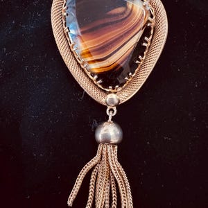 May include: A gold-toned brooch featuring a triangular, faceted brown and cream banded agate stone set in a textured metal frame. A polished gold-toned bead connects the main setting to a dangling tassel made of multiple fine gold-toned chains.