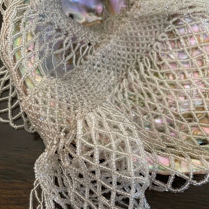 May include: A white beaded net draped over a large iridescent seashell. The net is made of small, round beads that are woven together in a delicate pattern.