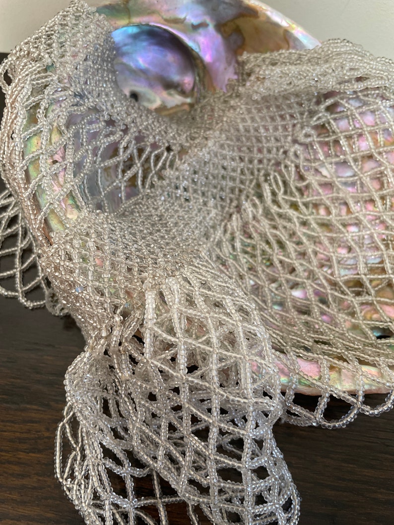 May include: A white beaded net draped over a large iridescent seashell. The net is made of small, round beads that are woven together in a delicate pattern.