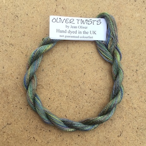 Könnte beinhalten: Ein Knäuel handgefärbter Stickgarn in Grün-, Blau- und Brauntönen. Das Garn ist kreisförmig angeordnet und mit dem Etikett "OLIVER TWISTS by Jean Oliver Hand dyed in the UK not guaranteed colourfast" versehen.