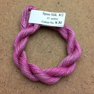 Puede incluir: Un ovillo de hilo de seda hilada en tonos rosas y morados, retorcido en forma circular. Una etiqueta blanca dice "Spun Silk 8/2, 15 metros, Colour No. S 31". El hilo se muestra sobre una superficie marrón.
