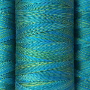 Op de afbeelding: Drie spoelen garen, elk met een blauwe en groene kleurverloop. Het garen is strak om de spoelen gewikkeld, waardoor een gestreept patroon ontstaat.