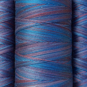 Puede incluir: Tres carretes de hilo con un esquema de color azul, morado y rojo. El hilo está enrollado apretado y tiene una apariencia texturizada.