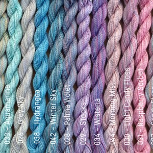 Puede incluir: Una colecci&oacute;n de 12 ovillos de hilo de bordar en varios tonos de azul, morado y rosa. El hilo est&aacute; dispuesto en filas, con cada ovillo etiquetado con un n&uacute;mero y un nombre de color.