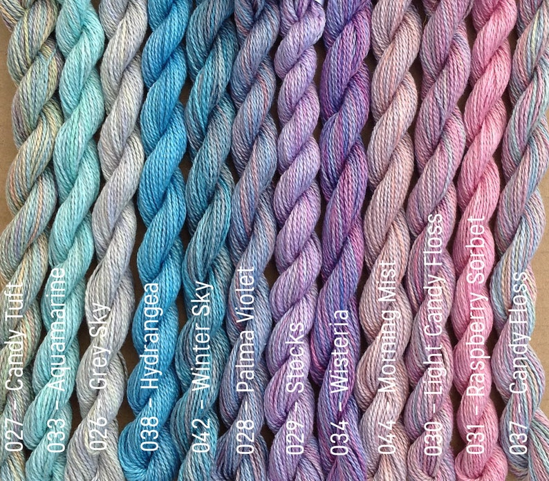 Puede incluir: Una colecci&oacute;n de 12 ovillos de hilo de bordar en varios tonos de azul, morado y rosa. El hilo est&aacute; dispuesto en filas, con cada ovillo etiquetado con un n&uacute;mero y un nombre de color.