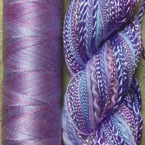 May include: A spool of purple and blue yarn with a skein of multi-colored yarn with silver thread. The skein is a mix of purple, blue, and pink.