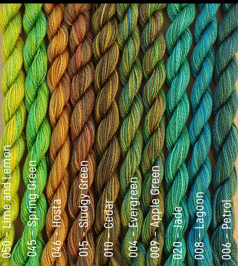 Puede incluir: Una colecci&oacute;n de diez ovillos de hilo te&ntilde;ido a mano en tonos de verde, amarillo y azul. El hilo est&aacute; dispuesto en una fila, con los ovillos etiquetados con n&uacute;meros y nombres.