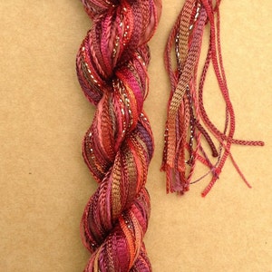 May include: A skein of hand-dyed silk ribbon in shades of red, orange, and brown with silver metallic thread. The label reads "OLIVER TWIST by Jean Oliver Hand dyed in the UK not guaranteed colorfast".