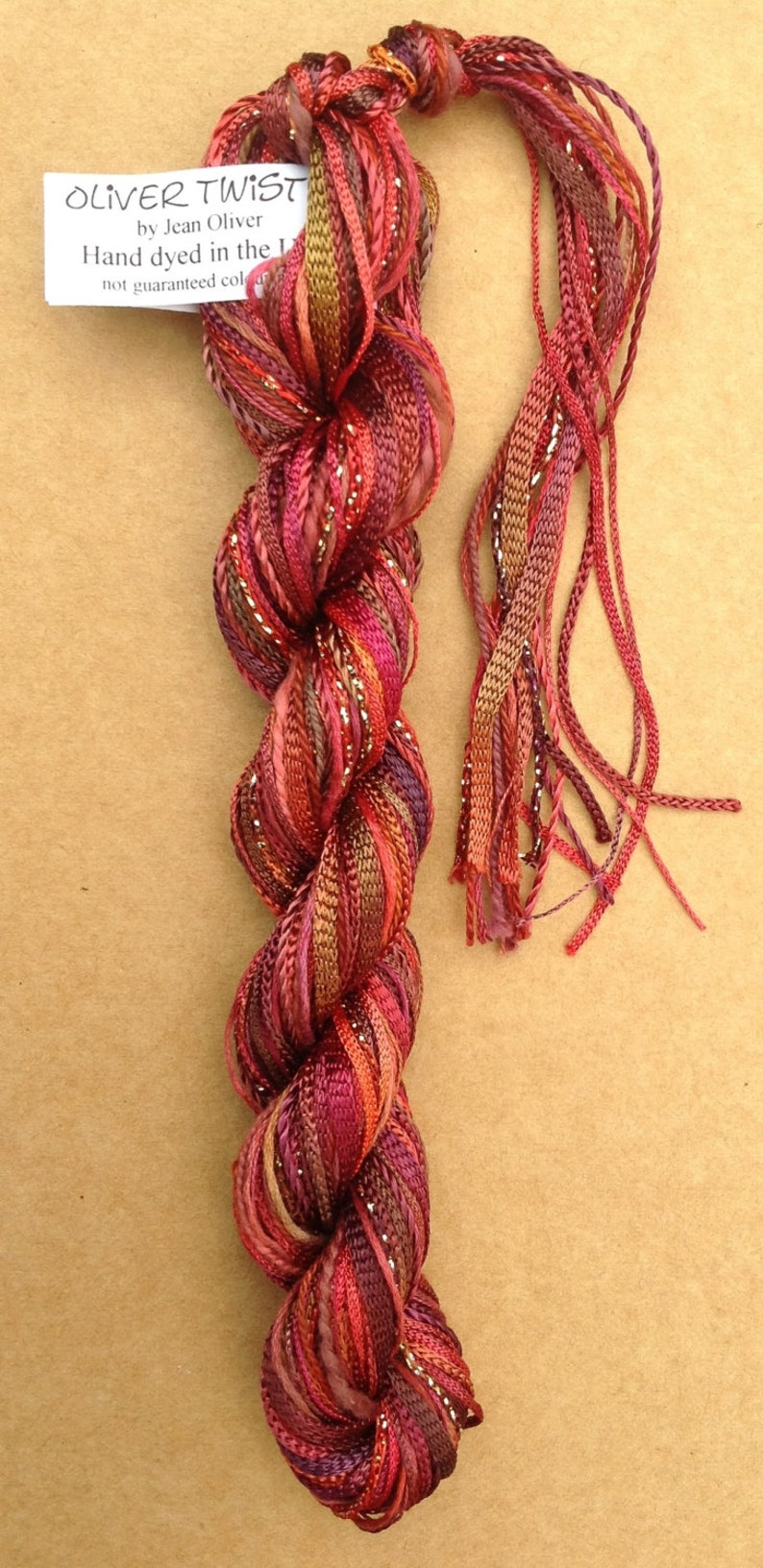 May include: A skein of hand-dyed silk ribbon in shades of red, orange, and brown with silver metallic thread. The label reads "OLIVER TWIST by Jean Oliver Hand dyed in the UK not guaranteed colorfast".
