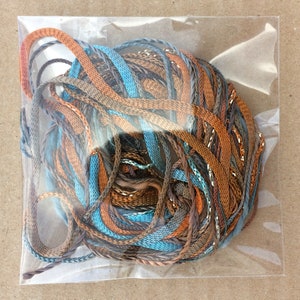May include: A close-up of a bundle of colourful embroidery floss in shades of brown, blue, and gold. The floss is wrapped in a clear plastic bag.