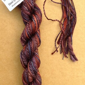 May include: A skein of hand-dyed yarn in shades of purple, brown, and orange with silver metallic thread. The yarn is labeled "Oliver Twists by Jean Oliver Hand dyed in the UK not guaranteed colourfast".