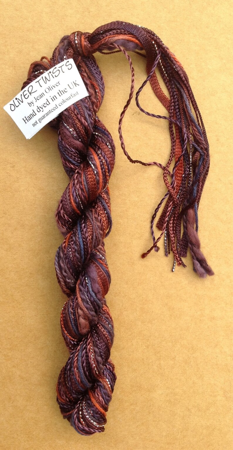 May include: A skein of hand-dyed yarn in shades of purple, brown, and orange with silver metallic thread. The yarn is labeled "Oliver Twists by Jean Oliver Hand dyed in the UK not guaranteed colourfast".