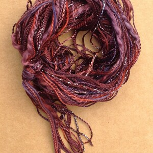 May include: A bundle of maroon, purple, and red yarn with silver thread woven throughout. The yarn is twisted into a circle and has long strands hanging from the bottom.