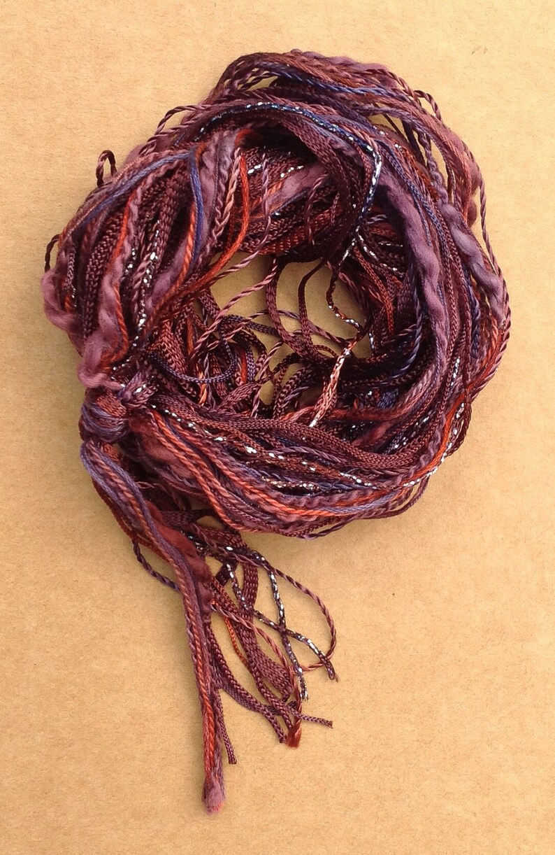 May include: A bundle of maroon, purple, and red yarn with silver thread woven throughout. The yarn is twisted into a circle and has long strands hanging from the bottom.
