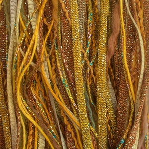 May include: A close-up of a bundle of colorful yarn strands in shades of gold, brown, and beige. Some strands have a sparkly finish.