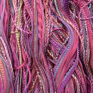 May include: A close-up of a bundle of colorful embroidery floss in shades of pink, purple, and brown. The floss is arranged in a random pattern, creating a textured and visually appealing display.