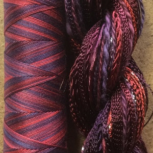 May include: Two spools of thread, one is a red and purple striped spool, the other is a loose bundle of red, purple, and silver thread.