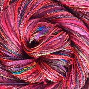 May include: A close-up shot of a colourful ball of yarn, predominantly in shades of red, pink, and purple. The yarn features a variety of textures, including ribbons, braided sections, and threads with metallic highlights. The yarn is tightly wound, creating a rich, textured appearance.