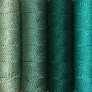 May include: A selection of thread spools in a range of greens and blues. The colours vary from pale seafoam to deep teal and turquoise. The thread is tightly wound on cylindrical spools, creating a gradient effect.