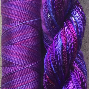 May include: A spool of purple and blue yarn with a strand of iridescent purple and blue yarn with silver glitter.
