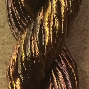 May include: A close-up of a skein of iridescent yarn with shades of brown, gold, and purple. The yarn is twisted and textured, creating a unique and eye-catching look.