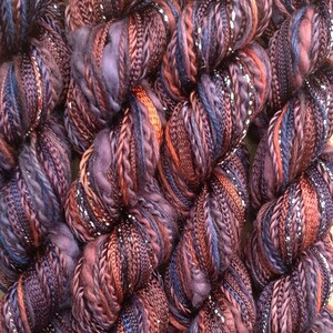 May include: Close-up of a bundle of yarn with a mix of purple, brown, and silver threads. The yarn is soft and fluffy, and it has a subtle sheen.